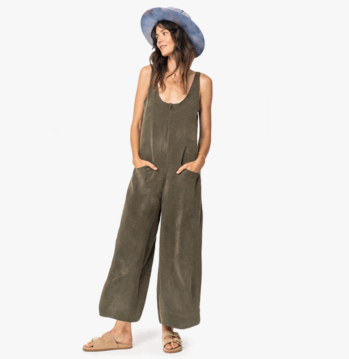 Person wearing a green mirage jumpsuit, wide-brimmed hat, and sandals, standing with hands in pockets.